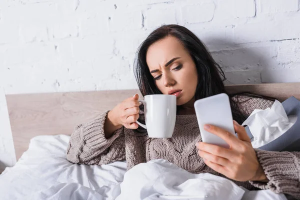 Malato giovane bruna donna con caldo bevanda e tessuto prendendo selfie a letto — Foto stock