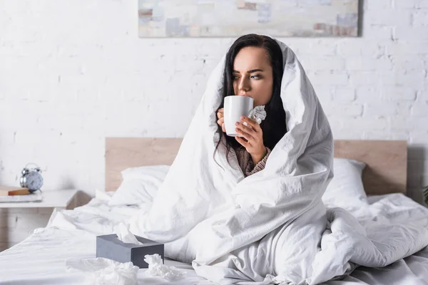 Kranke junge brünette Frau mit Heißgetränk und Gewebe im Bett — Stockfoto