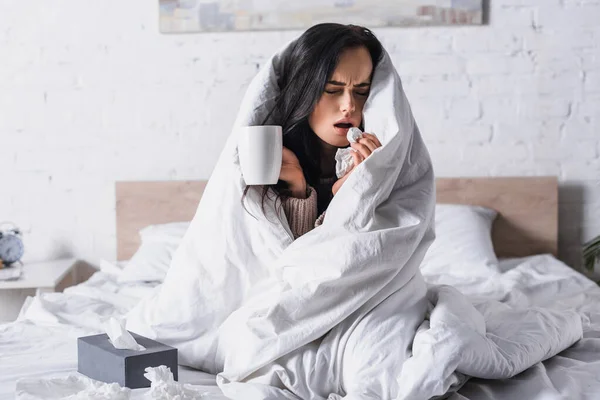 Sick young brunette woman with hot drink sneezing in bed — Stock Photo