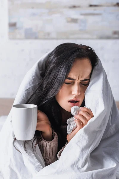 Kranke junge brünette Frau mit Heißgetränk niest im Bett — Stockfoto