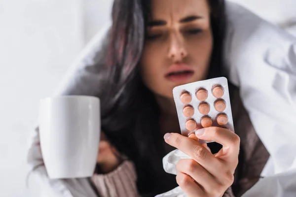 Sick young brunette woman with hot drink and pills in bed, blurred background — Stock Photo