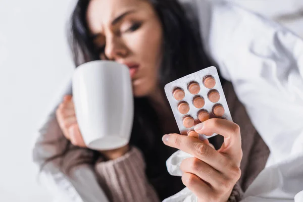 Kranke junge brünette Frau mit Heißgetränk und Tabletten im Bett, verschwommener Hintergrund — Stockfoto