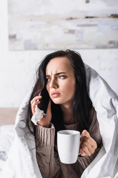 Malade jeune femme brune avec boisson chaude et tissu parler sur smartphone au lit — Photo de stock