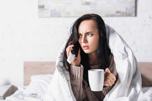 Kranke junge brünette Frau mit Heißgetränk und Gewebegesprächen auf Smartphone im Bett — Stockfoto