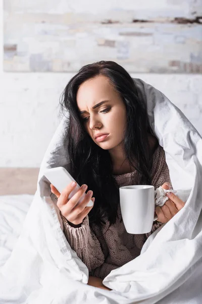 Kranke junge brünette Frau mit Heißgetränk und Gewebe mit Smartphone im Bett — Stockfoto