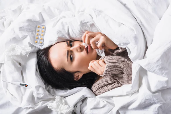 Top view of sick young brunette woman with tissue, pills and thermometer  in bed — Stock Photo