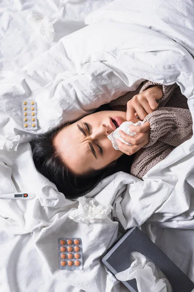 Draufsicht auf kranke junge brünette Frau mit Gewebe, Tabletten und Thermometer im Bett — Stockfoto