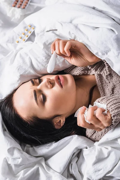 Top view of sick young brunette woman with tissue and thermometer in bed — стоковое фото