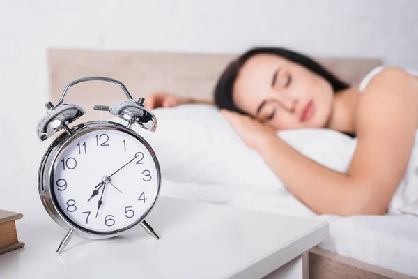 Класичний будильник і розмита молода брюнетка жінка спить у ліжку на фоні — Stock Photo