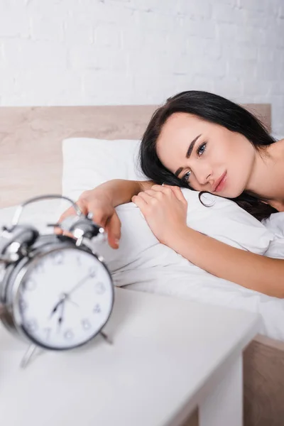 Verschwommene klassische Wecker und junge brünette Frau im Bett auf dem Hintergrund — Stockfoto