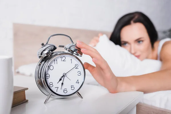 Klassischer Wecker und verschwommene junge brünette Frau im Bett auf dem Hintergrund — Stockfoto