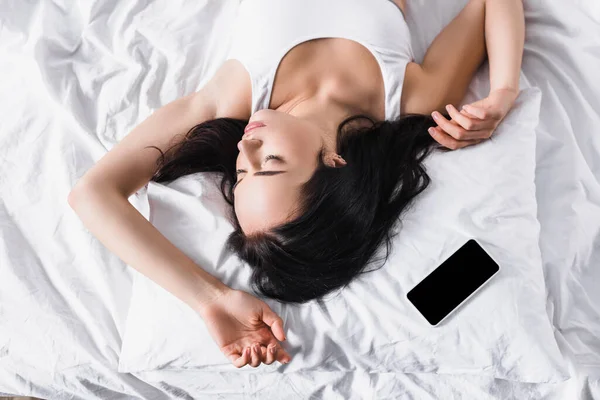 Вид зверху на молоду брюнетку, яка спить у ліжку біля смартфона — стокове фото