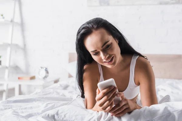 Lächelnde junge brünette Frau im Bett mit Smartphone — Stockfoto