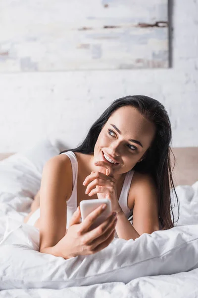 Soñadora sonriente joven morena en la cama usando smartphone - foto de stock