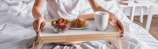 Cropped view of young woman having croissant and strawberry for breakfast in bed, banner — Stock Photo