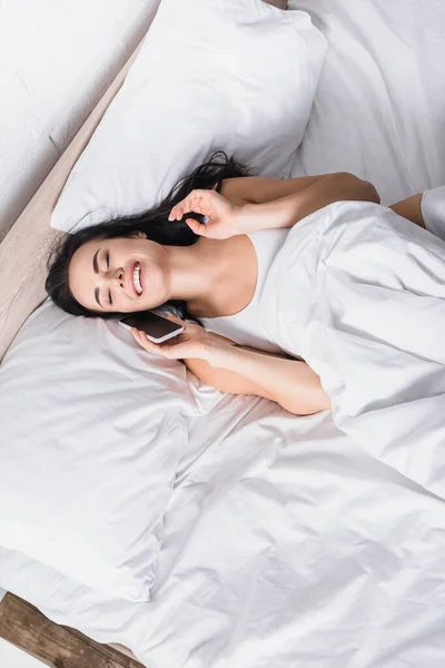 Draufsicht auf glückliche junge brünette Frau, die auf Smartphone im Bett spricht — Stockfoto