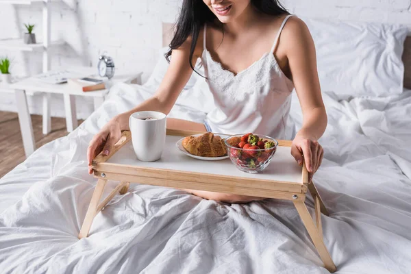 Vista cortada da jovem morena que tem croissant e morango para o café da manhã na cama — Fotografia de Stock