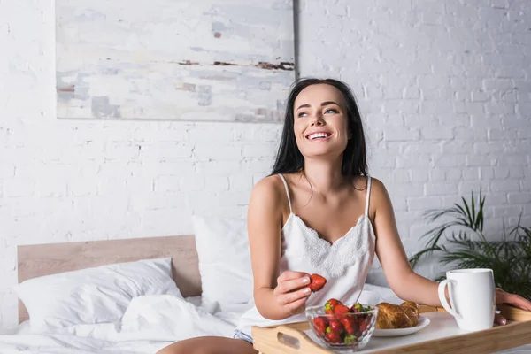 Счастливая молодая брюнетка с круассаном и клубникой на завтрак в постели — стоковое фото