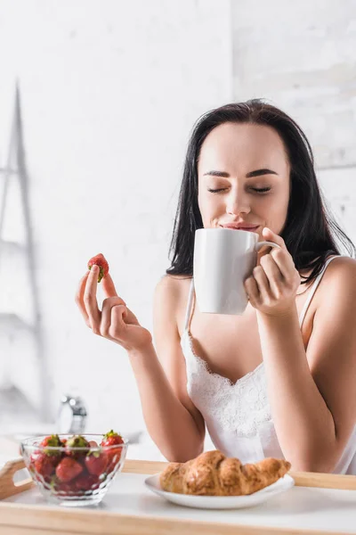 Jeune femme brune ayant croissant et fraise pour le petit déjeuner et boire du cacao au lit — Photo de stock