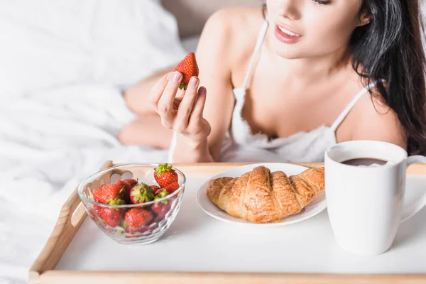 Cropped view of young brunette woman having croissant and strawberry for breakfast and drinking cocoa in bed — Stock Photo