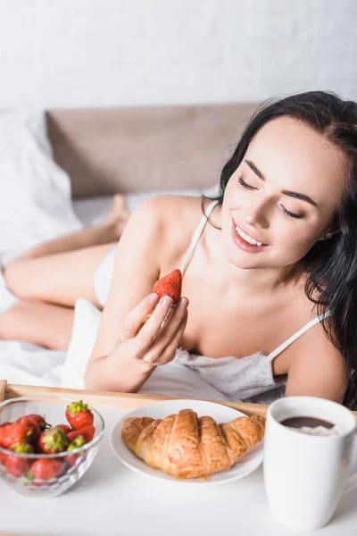Giovane donna bruna con croissant, fragola e cacao per la colazione a letto — Foto stock