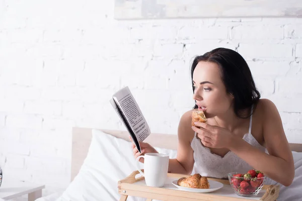 Junge brünette Frau frühstückt mit Croissant, Erdbeere und Kakao, während sie im Bett Zeitung liest — Stockfoto