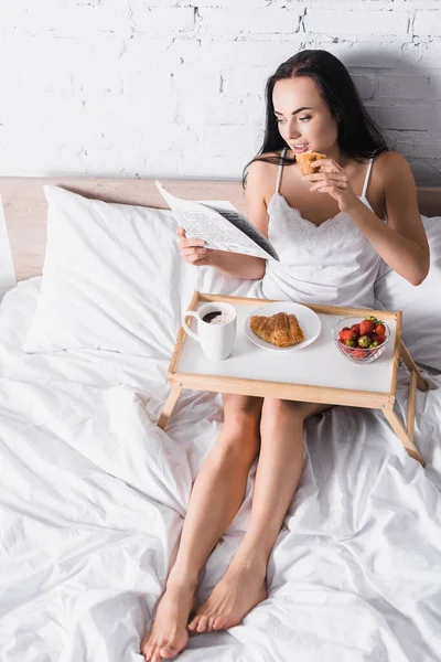 Junge brünette Frau frühstückt mit Croissant, Erdbeere und Kakao, während sie im Bett Zeitung liest — Stockfoto