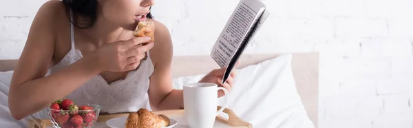 Vista recortada de la mujer morena tomando croissant, fresa y cacao para el desayuno mientras lee el periódico en la cama, pancarta - foto de stock