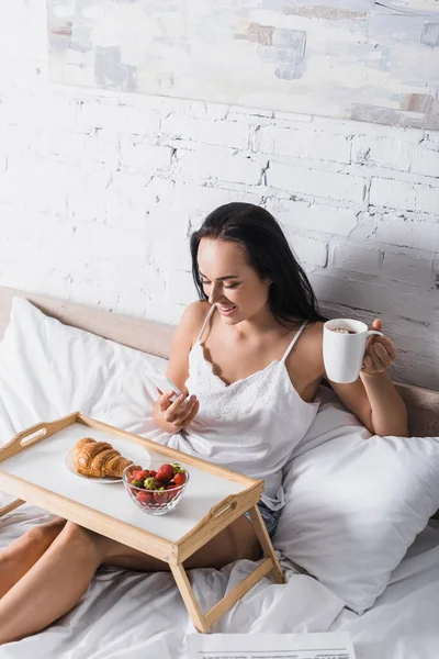 Jovem morena tendo croissant, morango e cacau para o café da manhã enquanto usa smartphone na cama — Fotografia de Stock