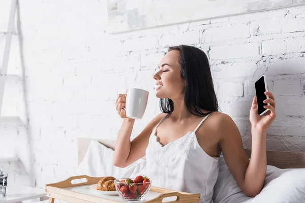 Young brunette woman having croissant, strawberry and cocoa for breakfast while holding smartphone in bed — Stock Photo