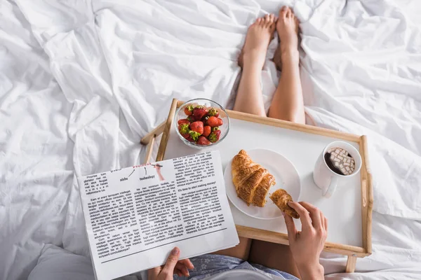 Cropped view of woman having croissant, strawberry and cocoa for breakfast while reading newspaper in bed — Stock Photo
