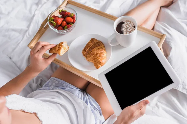 Vista ritagliata della donna che ha croissant, fragola e cacao per la prima colazione mentre tiene tablet digitale a letto — Foto stock