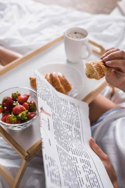 Cropped view of woman having croissant, strawberry and cocoa for breakfast while reading newspaper in bed — Stock Photo