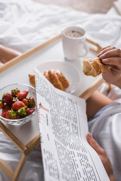 Vista recortada de la mujer que toma croissant, fresa y cacao para el desayuno mientras lee el periódico en la cama - foto de stock