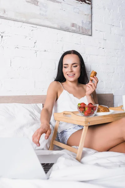 Lächelnde junge brünette Frau frühstückt im Bett, während sie Laptop benutzt — Stockfoto