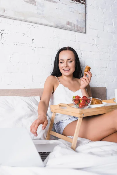 Sorrindo jovem morena mulher tomando café da manhã na cama enquanto usa laptop — Fotografia de Stock