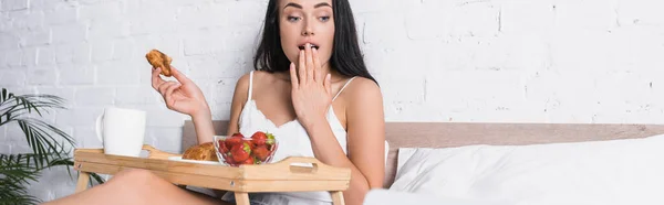 Giovane donna bruna scioccata con croissant, fragola e cacao per la colazione a letto, banner — Foto stock