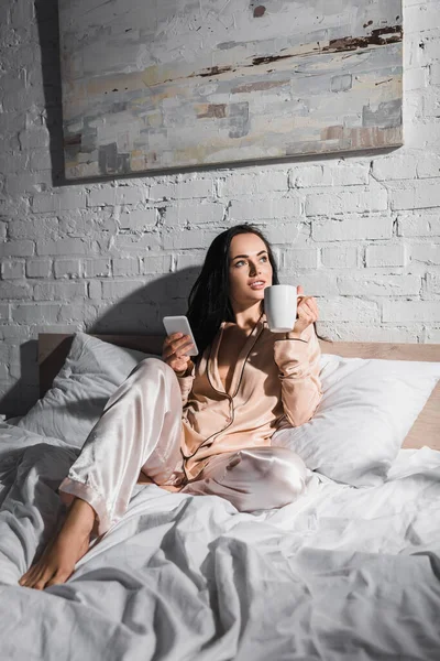 Junge brünette Frau sitzt morgens mit Becher und Smartphone im Bett — Stockfoto