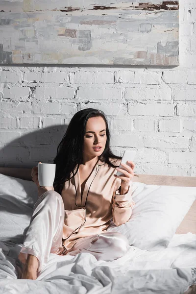 Jeune femme brune assise au lit avec tasse et smartphone le matin — Photo de stock