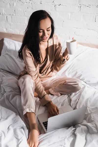 Giovane donna bruna seduta a letto con tazza e laptop al mattino — Foto stock