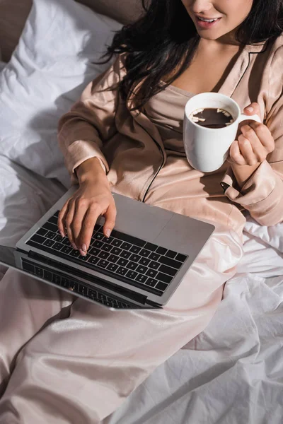 Vista cortada de jovem morena sentada na cama com caneca e laptop pela manhã — Fotografia de Stock
