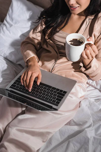 Cropped view of young brunette woman sitting in bed with mug and laptop at morning — Stock Photo