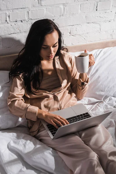 Jovem morena sentada na cama com caneca e laptop de manhã — Fotografia de Stock