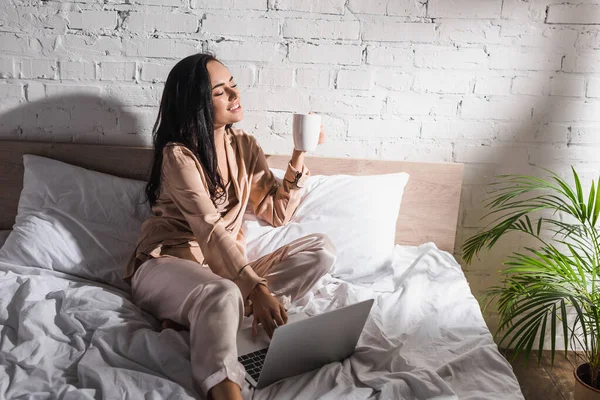 Улыбающаяся юная брюнетка, сидящая в постели с кружкой и ноутбуком по утрам — стоковое фото