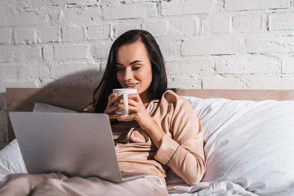 Jovem morena sentada na cama com caneca e laptop de manhã — Fotografia de Stock