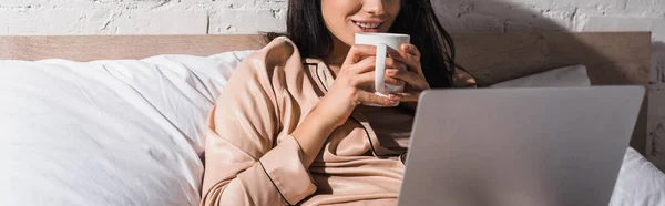 Vue recadrée de jeune femme brune assise au lit avec tasse et ordinateur portable le matin, bannière — Photo de stock