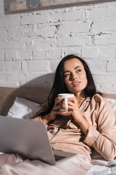 Jovem morena sentada na cama com caneca e laptop de manhã — Fotografia de Stock