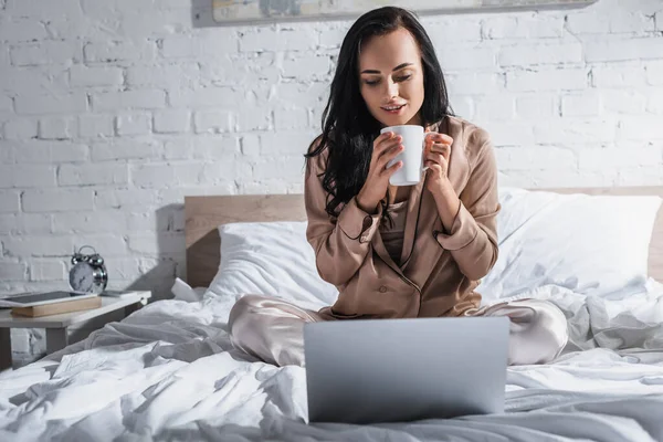 Jovem morena sentada na cama com caneca e laptop de manhã — Fotografia de Stock