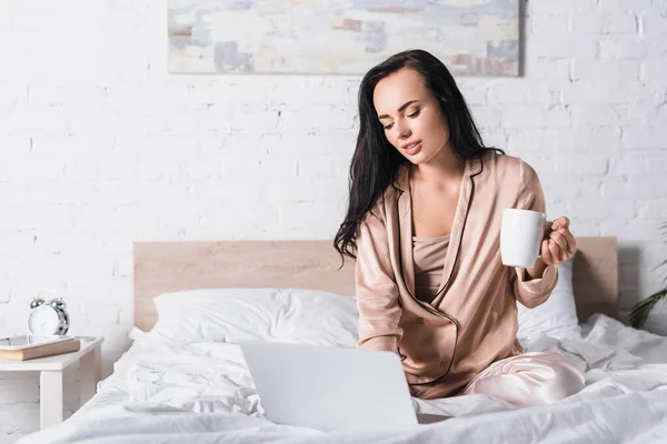 Young brunette woman sitting in bed with mug and laptop at morning — Stock Photo