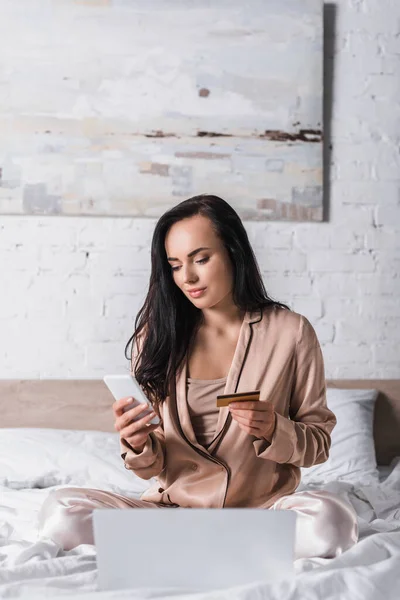 Junge brünette Frau sitzt morgens mit Smartphone, Kreditkarte und Laptop im Bett — Stockfoto